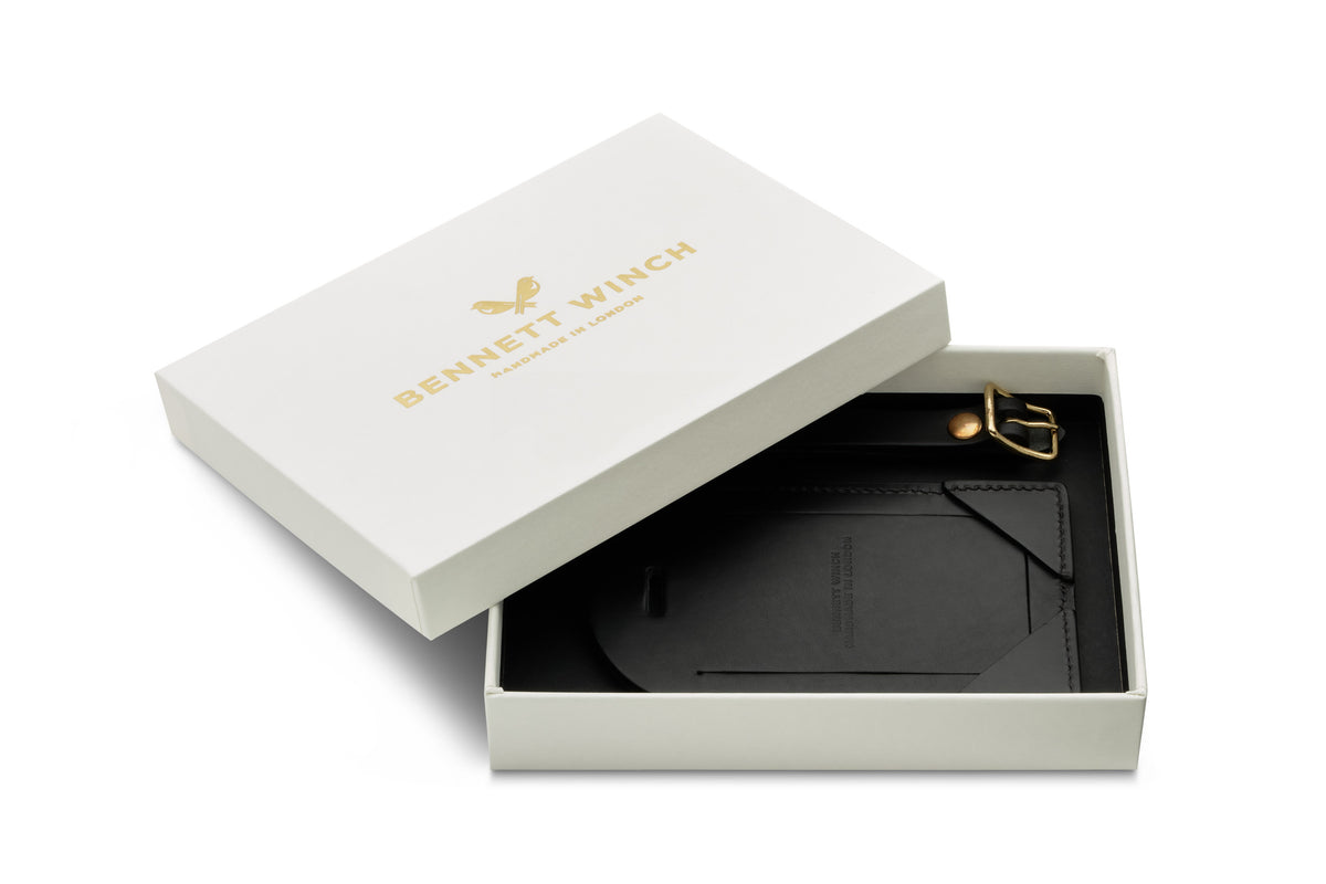 Open white Bennett Winch box, inside is a black leather luggage tag and leather strap with buckle.